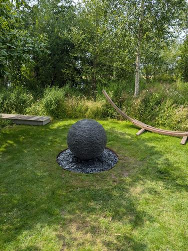 800mm (medium sized) slate ball water feature with a sump reservoir installed in the ground.The area is surround by lush green grass and bordered by wild flower medows.