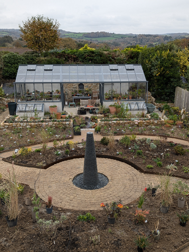 Tall Cornish chimney slate water feature, set in a circular central brick path. Located halfway between the house and the bottom of the garden, where a large green house takes pride..